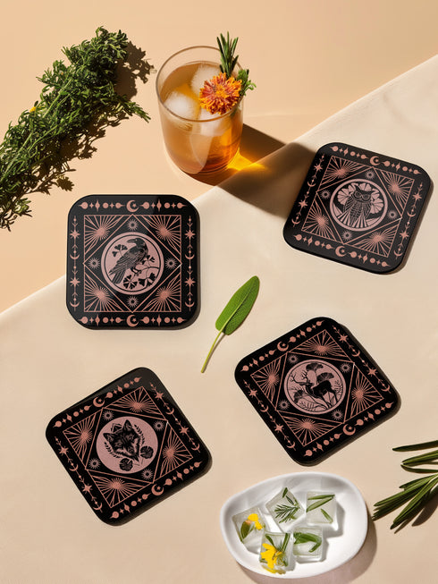 A set of four black, high-gloss square coasters featuring intricate rose-gold metallic illustrations of an owl, raven, stag, and wolf framed by celestial and botanical motifs. Made from durable masonite hardboard with cork backing for slip-resistant protection.